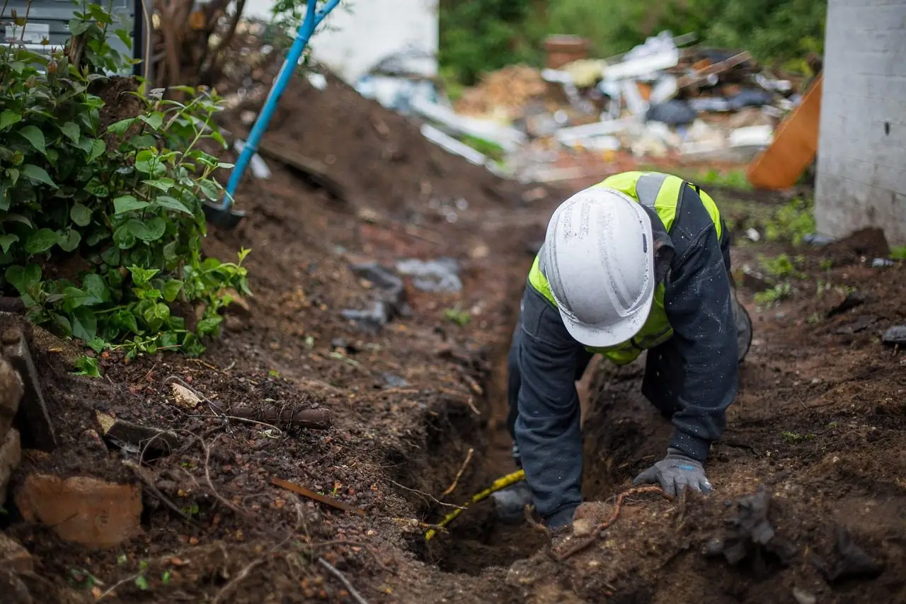 Worker in a hard hat kneeling in a narrow trench beside plants